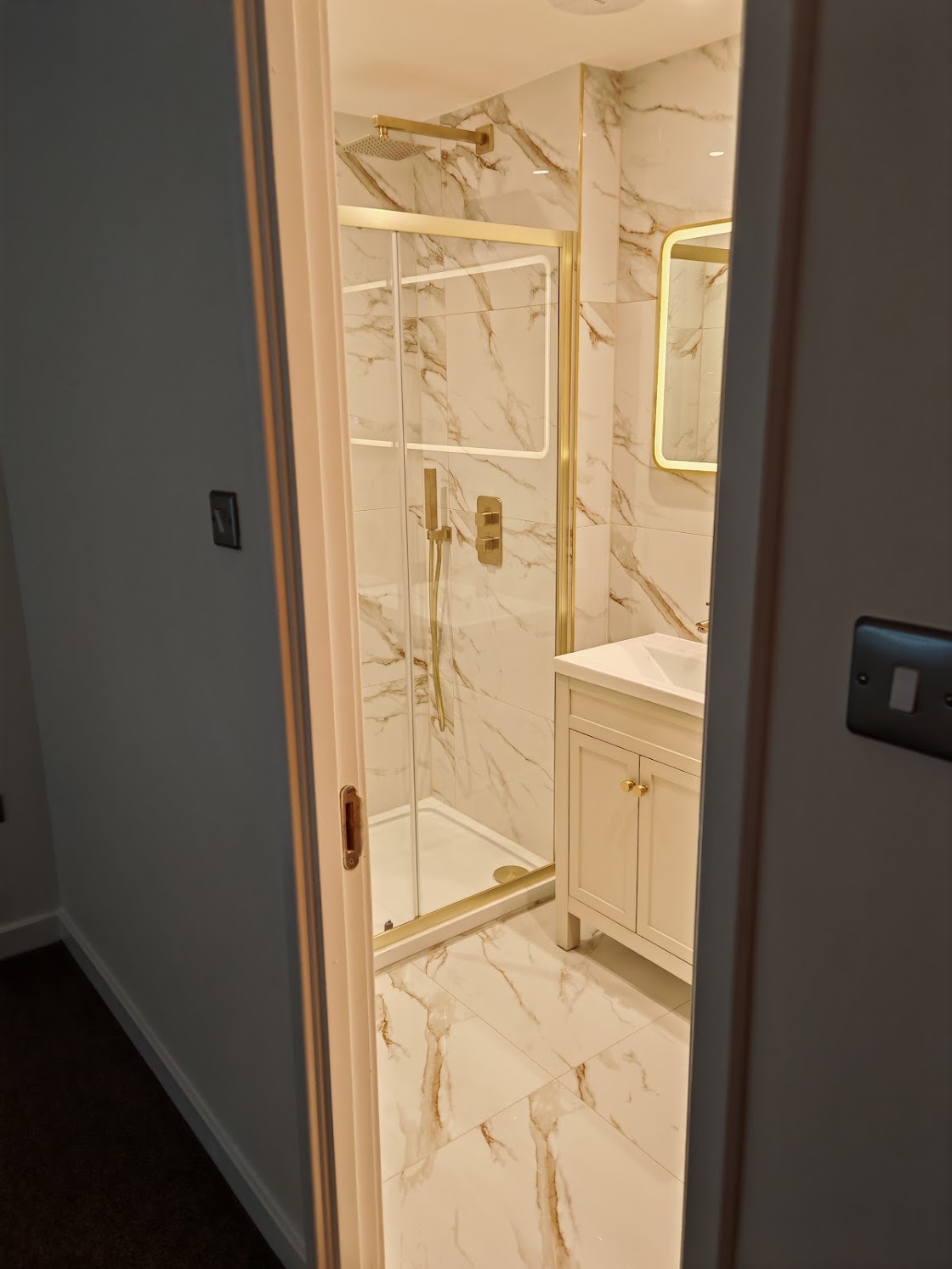 View through doorway into the en-suite showing gold-framed rain shower enclosure and LED backlit mirror